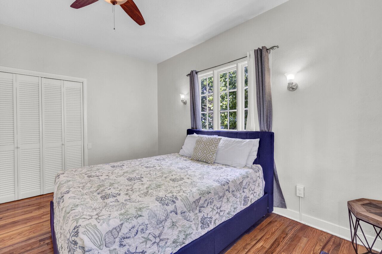 Une chambre à coucher avec un lit, une fenêtre avec des rideaux, un ventilateur de plafond et un plancher en bois.