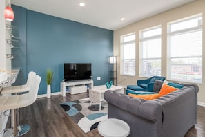 Ein modernes Wohnzimmer mit einem großen Flachbildfernseher, einem grauen Sofa mit orangenen Kissen, einem weißen Couchtisch und einer blauen Akzentwand.