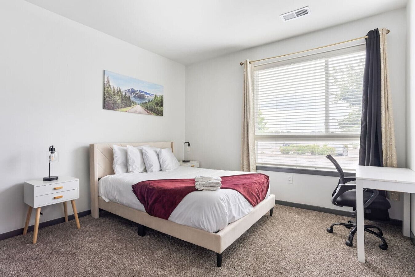 A bedroom with a bed, a desk, a chair, a nightstand, and a window with blinds.