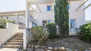 Un edificio con escalera de concreto, un pequeño jardín con arbustos y un árbol alto, y fachada blanca.