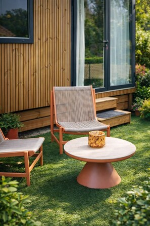 A modern outdoor seating area with a round table and two cushioned chairs, a wooden deck, and a glass door leading to a garden.