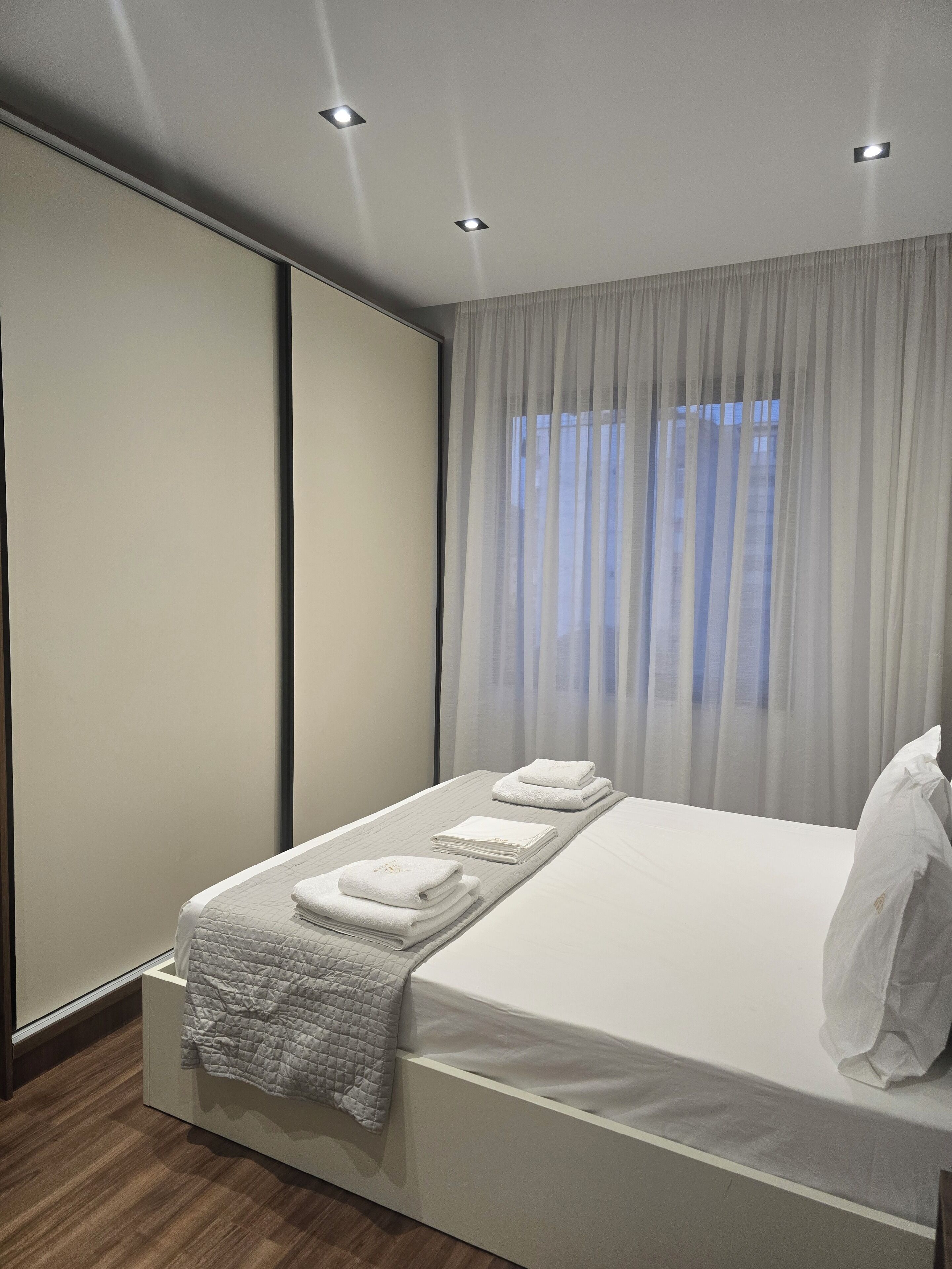 A modern hotel room with a neatly made bed, a large window with sheer curtains, and a wooden floor.