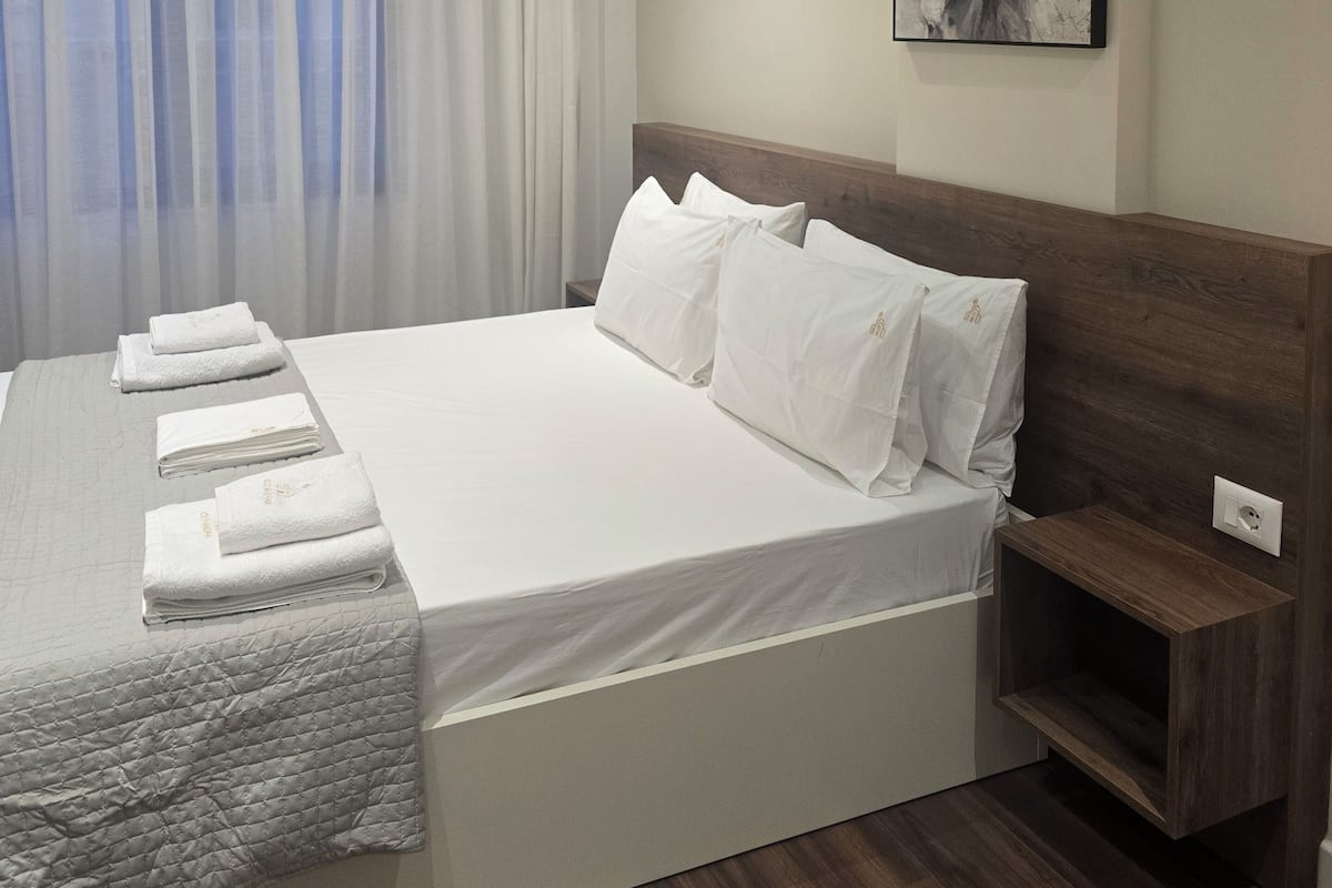 Ein modernes Hotelzimmer mit einem ordentlich bezogenen Bett, einem Nachttisch, einem Wandbild und einem großen Fenster mit durchsichtigen Vorhängen.