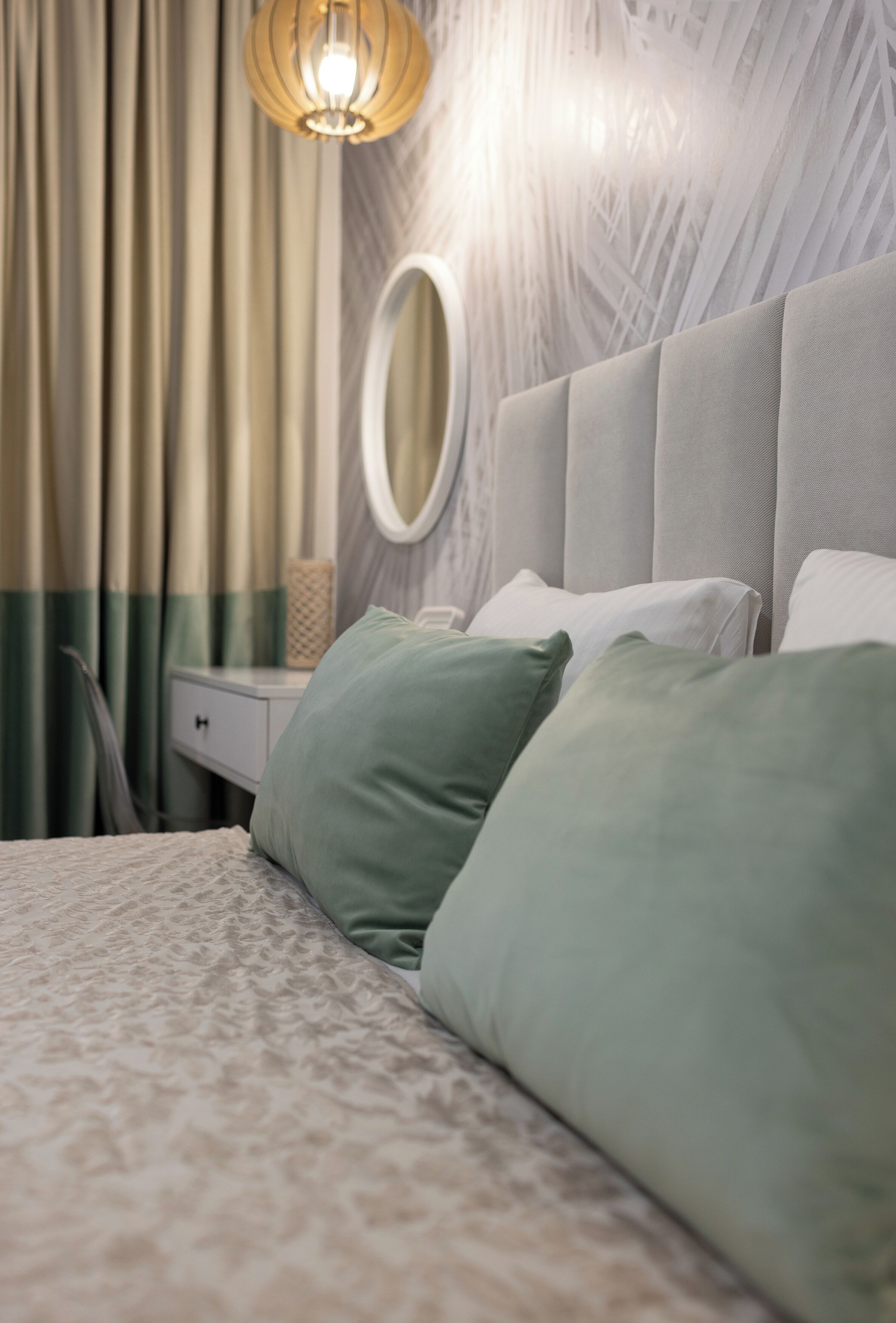 Een netjes opgemaakt bed met groene en witte kussens, een nachtkastje met een lamp en een spiegel aan de muur.