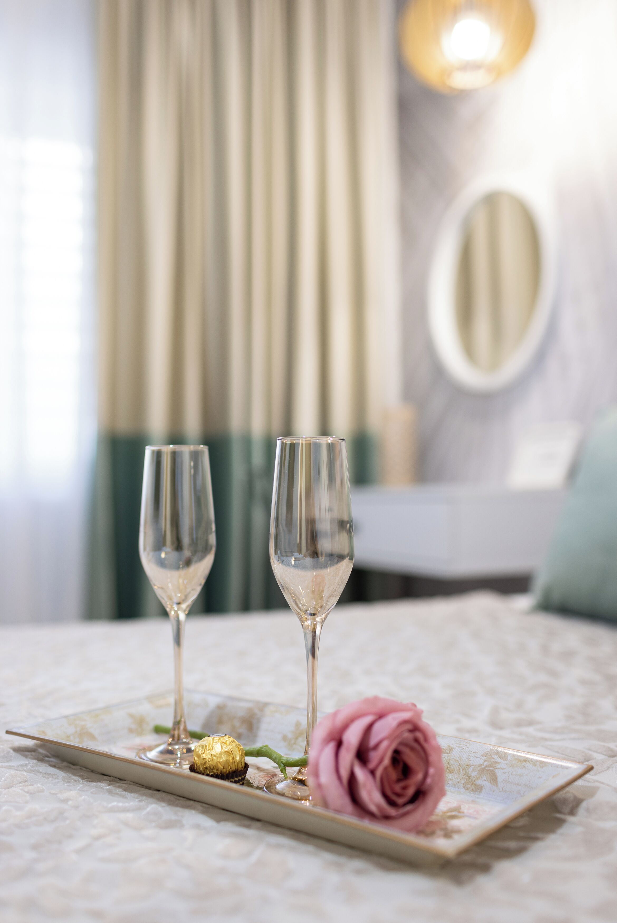 A table with two glasses of water, a pink rose, and a chocolate truffle on a decorative tray in a hotel room.