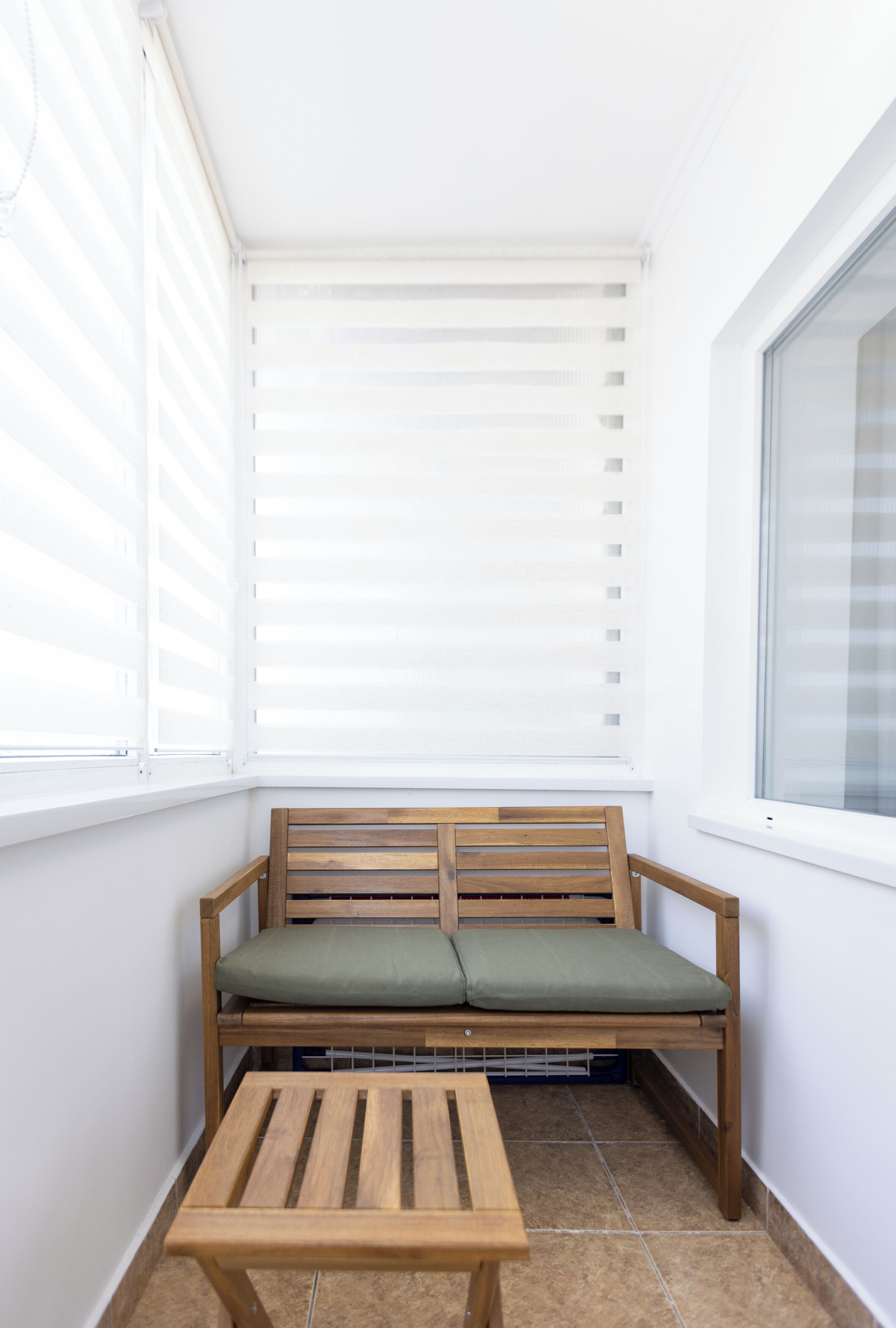 A small, enclosed balcony with a wooden bench and a table, featuring white walls and a window with blinds.