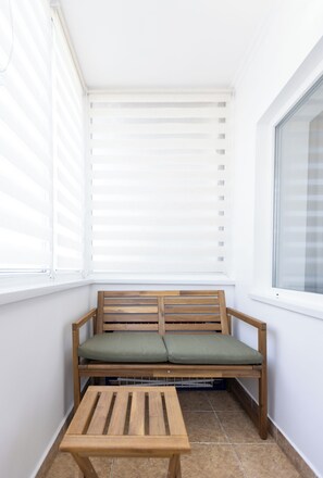 A small, enclosed balcony with a wooden bench and a table, featuring white walls and a window with blinds.