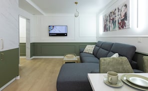 A modern living room with a grey sofa, a small table with a cup and plates, a television on the wall, and a painting above the sofa.