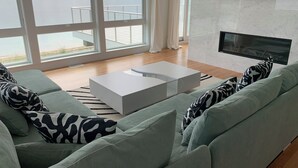 Una sala de estar moderna con un gran ventanal que da a un balcón y al mar. Hay una mesa de centro blanca y un sofá gris con cojines estampados.