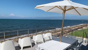 Una terraza con sillas de plástico blanco y una mesa, con vista a una playa y olas, y una costa lejana.