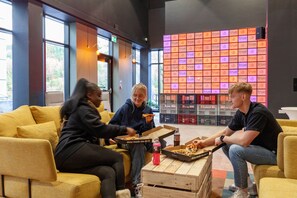 A group of people sitting on yellow sofas in a modern indoor space with large windows and a wall displaying a grid of colorful squares. - For Students Only True Swansea (Swansea)