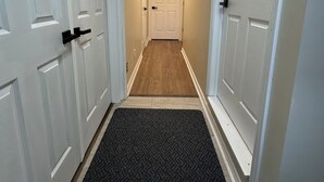 A narrow hallway with white doors on both sides, a wooden floor, and a black doormat at the entrance.