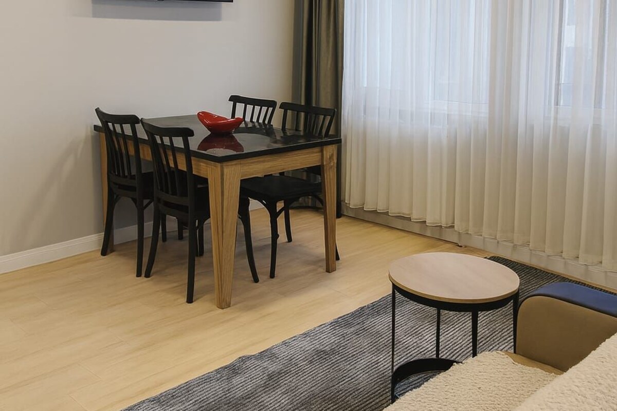 A modern room with a dining table, chairs, a sofa, a TV mounted on the wall, and a large window with curtains.