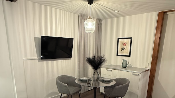 Ein modernes Wohnzimmer mit einer Couch, einem runden Esstisch mit Stühlen, einem an der Wand befestigten Fernseher und einer Küchenzeile mit Arbeitsplatte und Schränken.