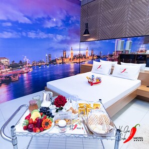 Una habitación de hotel con cama, un carrito de desayuno con frutas, bollería y una botella de jugo.