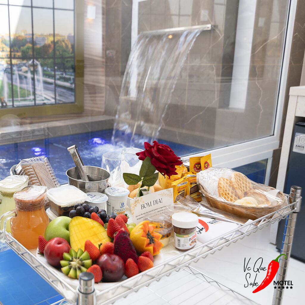 A breakfast buffet with various fruits, cheeses, and pastries, set against a backdrop of a water feature and a city view.