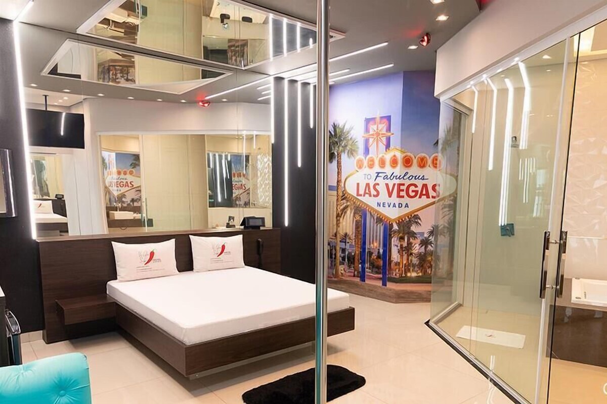Habitación de hotel con cama, escritorio, silla, puerta de vidrio y un letrero de neón que dice "Welcome to Fabulous Las Vegas Nevada".