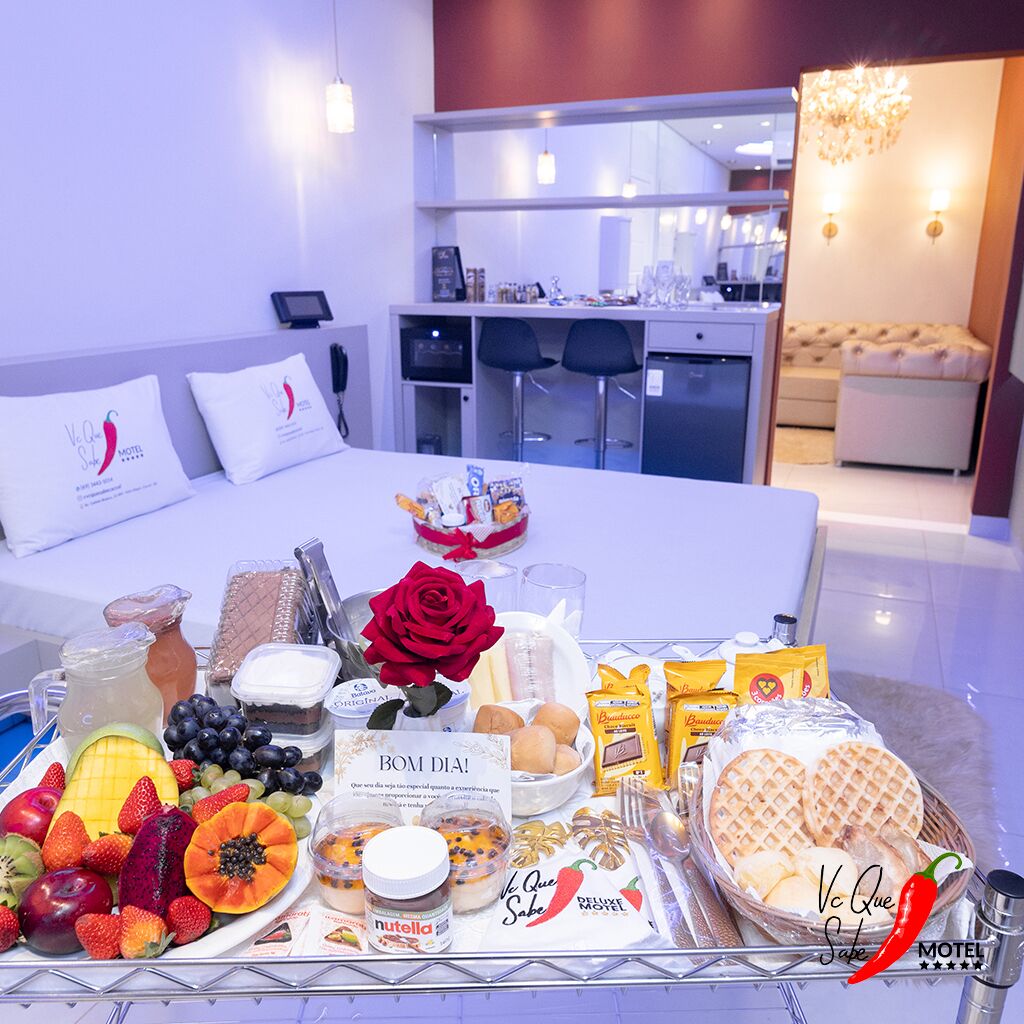 A hotel room with a neatly arranged breakfast tray on the bed, including fruits, pastries, and a red rose.