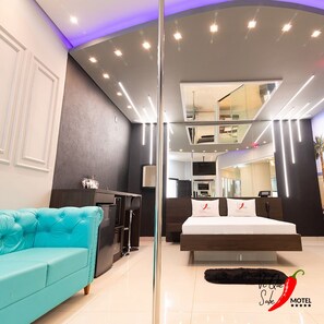 Una habitación de hotel moderna con un sofá color turquesa, una cama con cabecera blanca, una zona de cocina y un espejo grande.