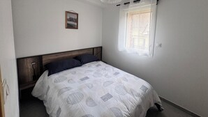 Een slaapkamer met een houten hoofdeinde, een bed met een wit beddegoed met een patroon, een hanglamp, een raam met dunne gordijnen en een ingelijst foto aan de muur.