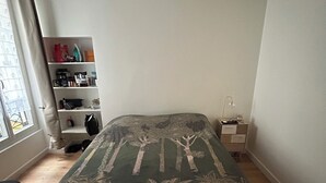 Un dormitorio con una cama, una mesita de noche, un estante con varios objetos y una ventana con cortinas.
