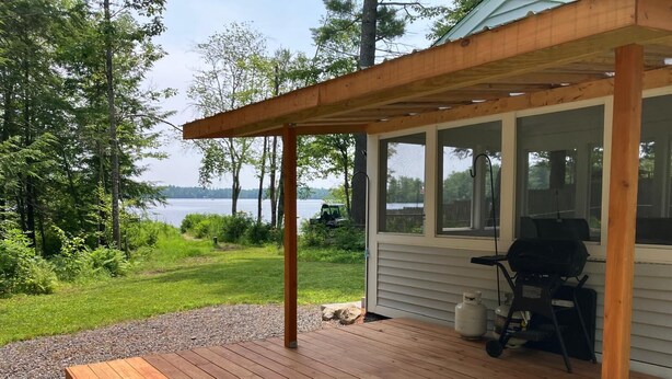 Un patio in legno con zona barbecue, affacciato su un lago con alberi sullo sfondo.
