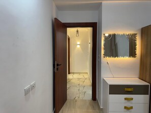 Um corredor com uma porta de madeira que dá para um quarto com piso de mármore. Há um armário branco com gavetas e um espelho com moldura singular.