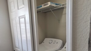 Ett tvättrum med en tvättmaskin och en torktumlare som man öppnar framifrån, en hylla med rengöringsmedel och en vit dörr.