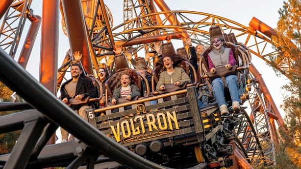 Una montaña rusa con el nombre 'Voltron' en la parte frontal, pasajeros sentados y una estructura metálica.