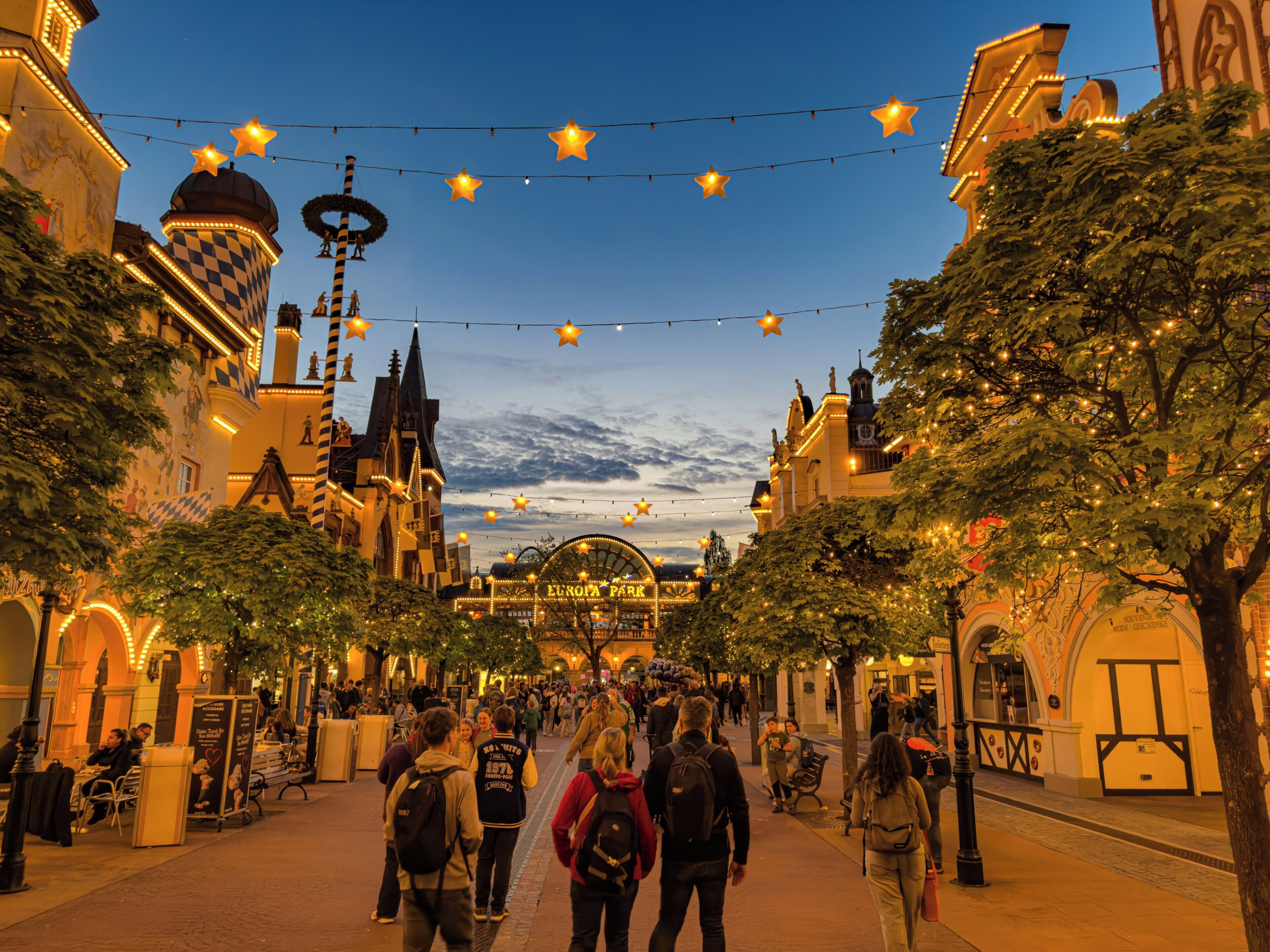 A street lined with trees and buildings, with people walking and a Ferris wheel in the background.