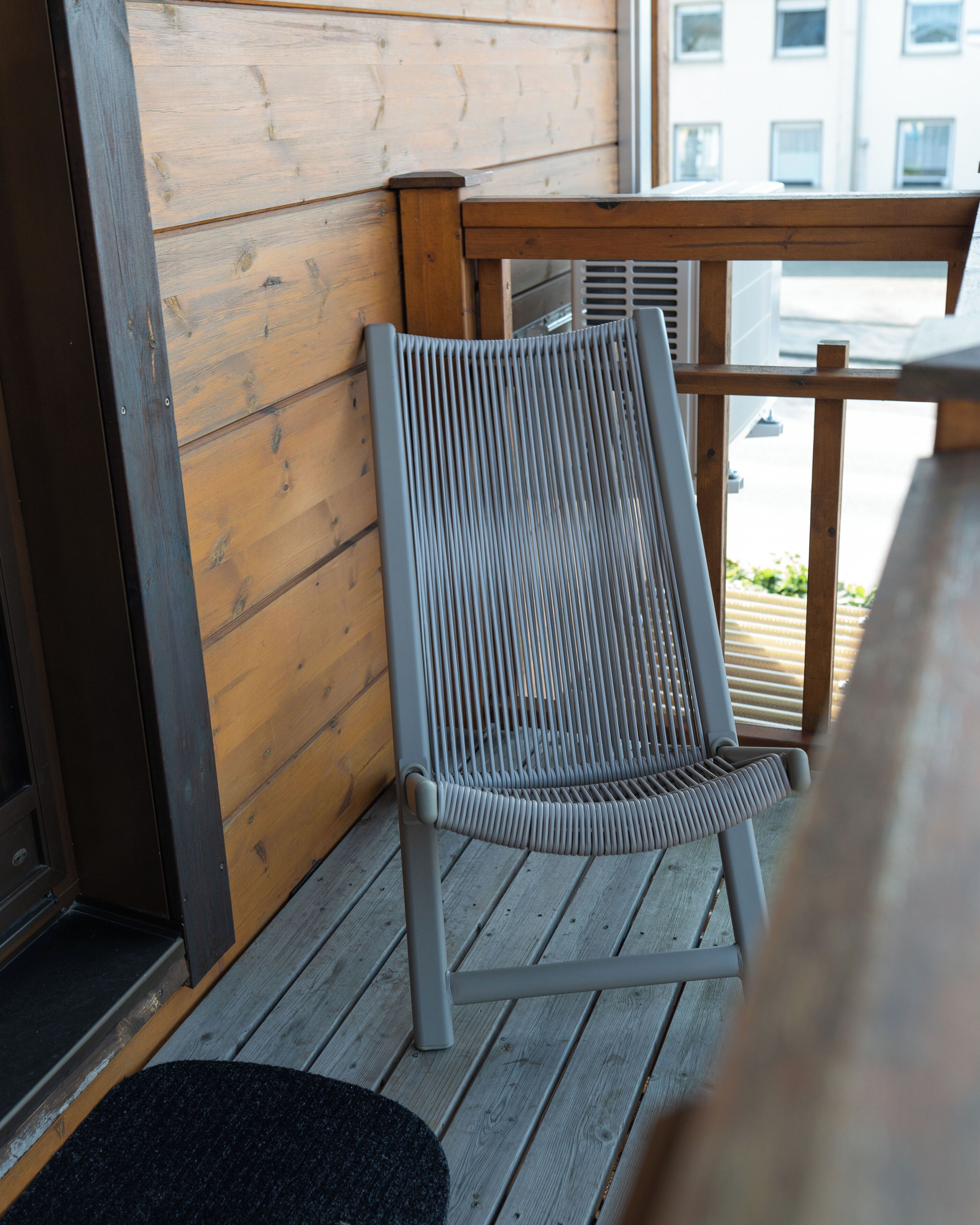 Une chaise avec un dossier à lattes, posée sur une terrasse en bois, un tapis noir et une fenêtre donnant sur des immeubles.