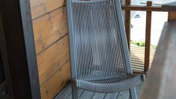 Une chaise avec un dossier à lattes, posée sur une terrasse en bois, un tapis noir et une fenêtre donnant sur des immeubles.