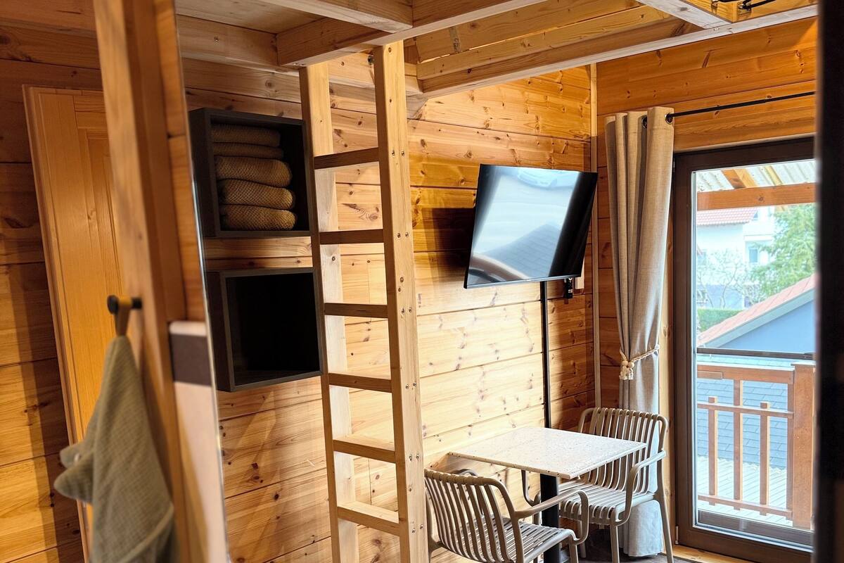 A wooden cabin interior with a TV, a table, chairs, a ladder, and a window.