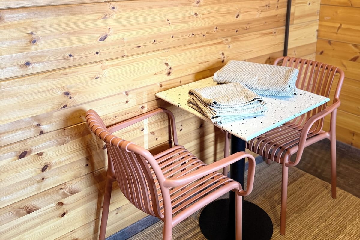A wooden cabin interior with a TV mounted on the wall, two wicker chairs, a table with a knitted blanket, and a framed picture of a bridge.