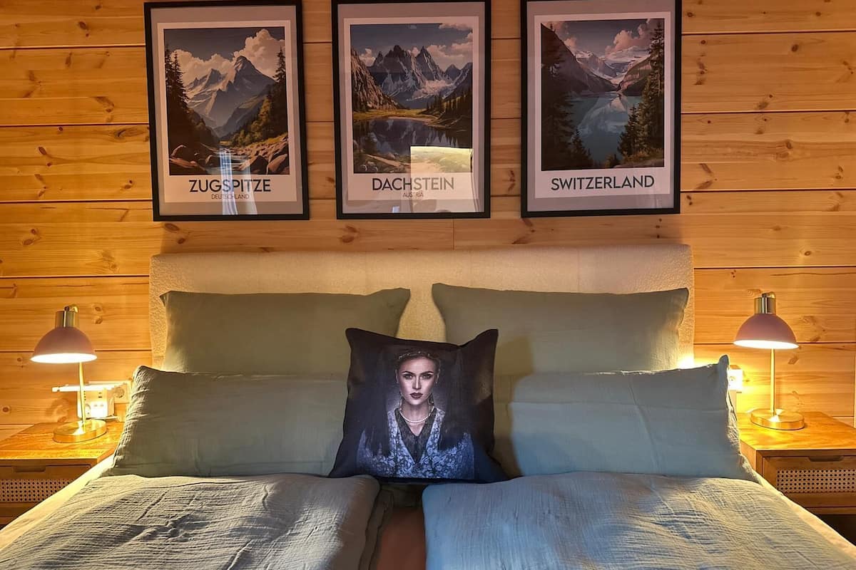 A wooden-paneled room with a bed, two bedside lamps, and three framed pictures of mountains labeled 'Zugspitze,' 'Dachstein,' and 'Switzerland.'