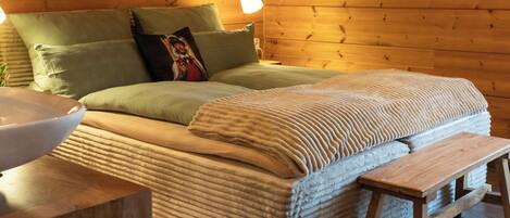 Interior de una cabaña de madera con cama, almohadas, mesita de noche y una planta colgante.