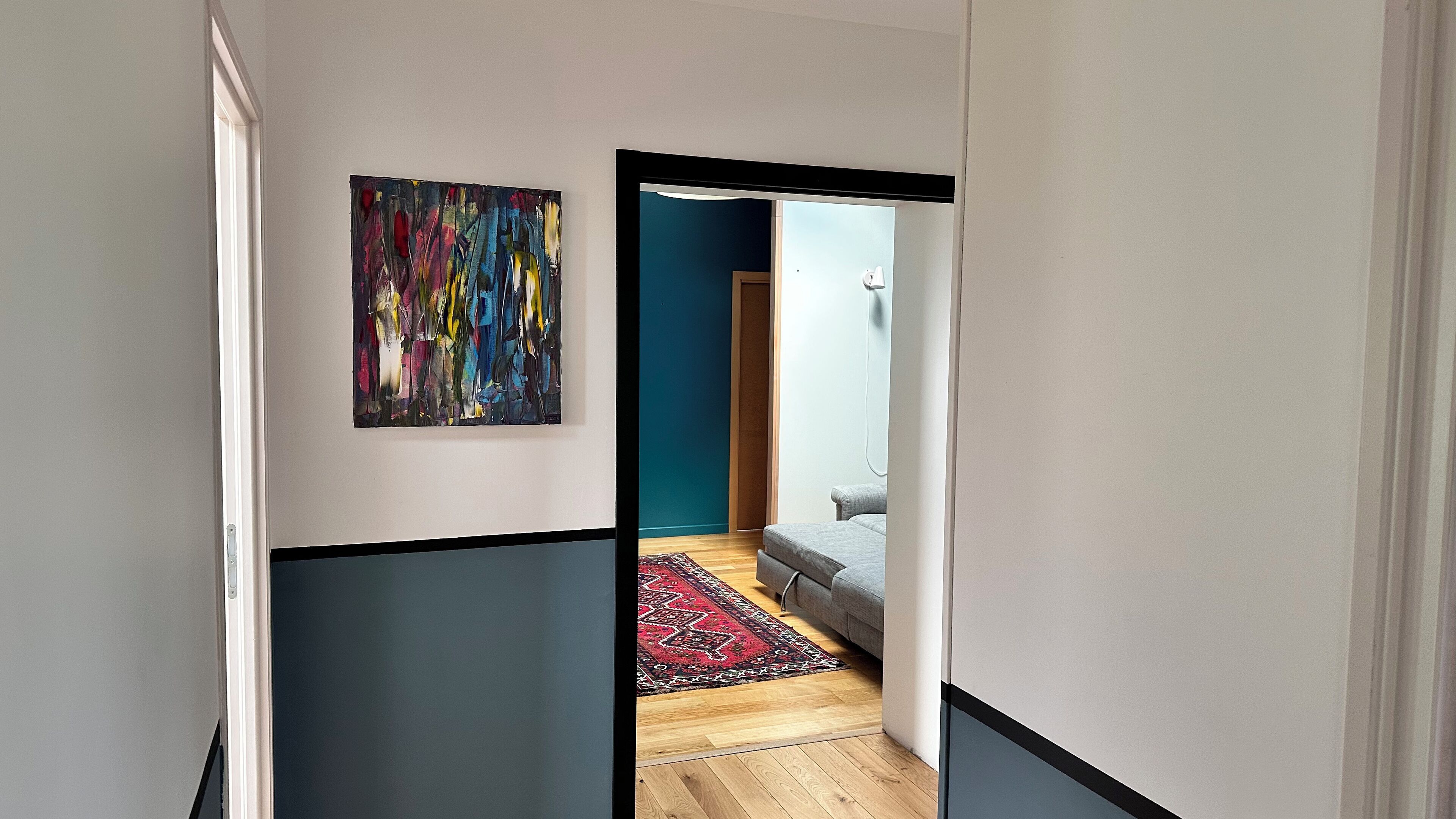 A hallway with a colorful abstract painting, a patterned rug, and a grey sofa in a room with teal walls.