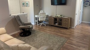 Een woonkamer met een beige bank, een fauteuil, een klein wit bureau, een aan de muur bevestigde televisie en een kast met decoratieve bloemen.