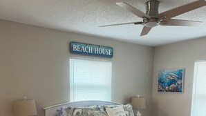 Een slaapkamer met een bed, nachtkastje, lamp, plafondventilator, vloerkleed en een bord aan de muur met de tekst 'BEACH HOUSE'.