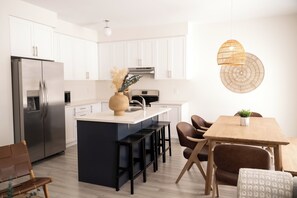 A modern kitchen with white cabinets, a stainless steel refrigerator, a central island with bar stools, and a wooden dining table with chairs.