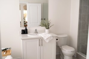 A modern bathroom with a white sink, mirror, and toilet. There is a potted plant on the sink counter and a towel hanging on the cabinet door. - Spacious 4BR Home|Groups|Families|Trail Access|WiFi|Parking (Quinte West)