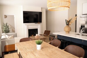 A modern living room with a fireplace, a dining table with chairs, a TV mounted on the wall, and a kitchen area with a sink and cabinets. - Spacious 4BR Home|Groups|Families|Trail Access|WiFi|Parking (Quinte West)