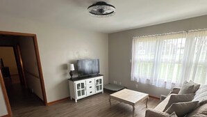 Ein Wohnzimmer mit Holzboden, einem weißen Schrank, einem Fernseher, einem beigen Sofa und einem Fenster mit durchsichtigen Vorhängen.