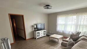 Ein geräumiges Wohnzimmer mit einer großen Couch, einem Couchtisch und einem Fernseher auf einem Schrank. Eine Tür führt in einen weiteren Raum, und ein Fenster mit Vorhängen ist ebenfalls vorhanden.