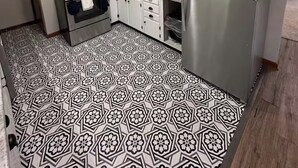 Eine kompakte Küche mit weißen Schränken, einem Edelstahl-Kühlschrank, einer Mikrowelle und einem Gasherd. Der Boden ist mit einem geometrischen Schwarz-Weiß-Fliesenmuster ausgelegt.