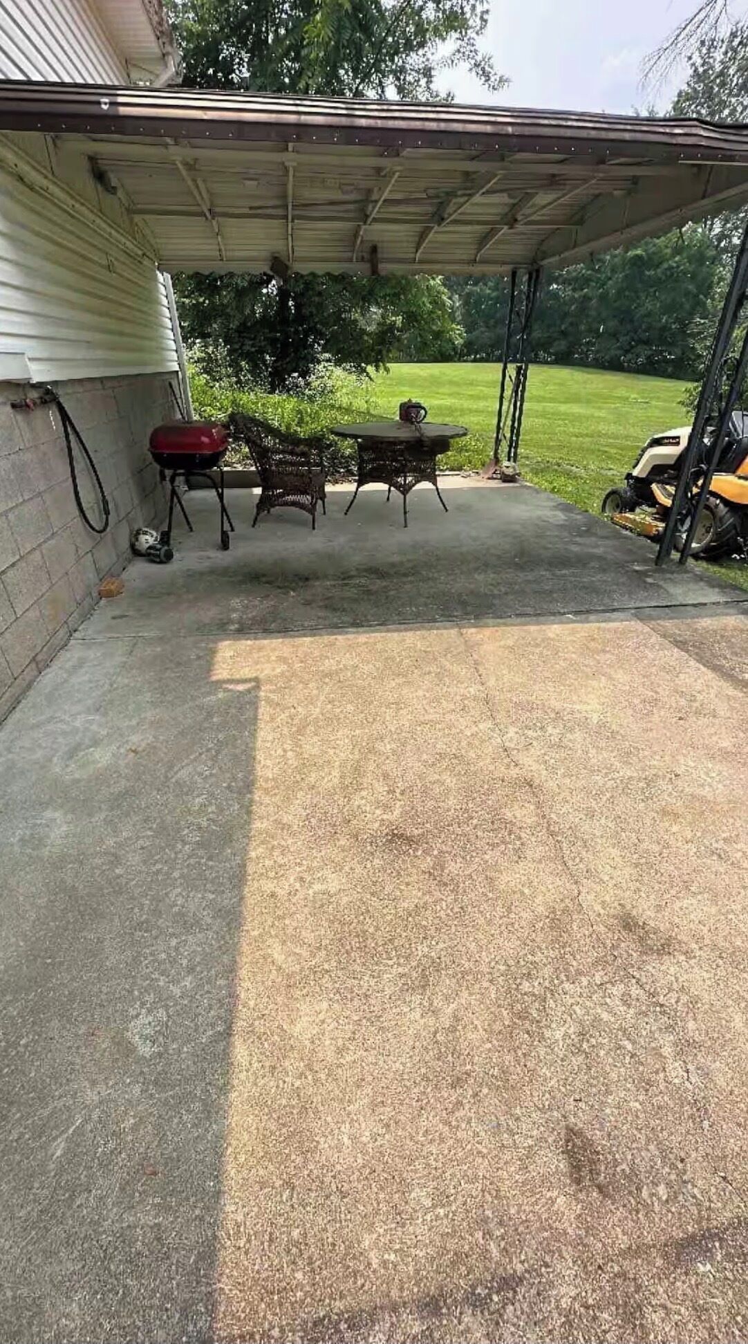 A covered patio area with a concrete floor, a table and chairs, a red grill, and a lawnmower in the background.
