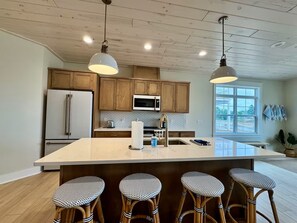 A modern kitchen with wooden cabinets, a large island with bar stools, a refrigerator, and a window with a view.