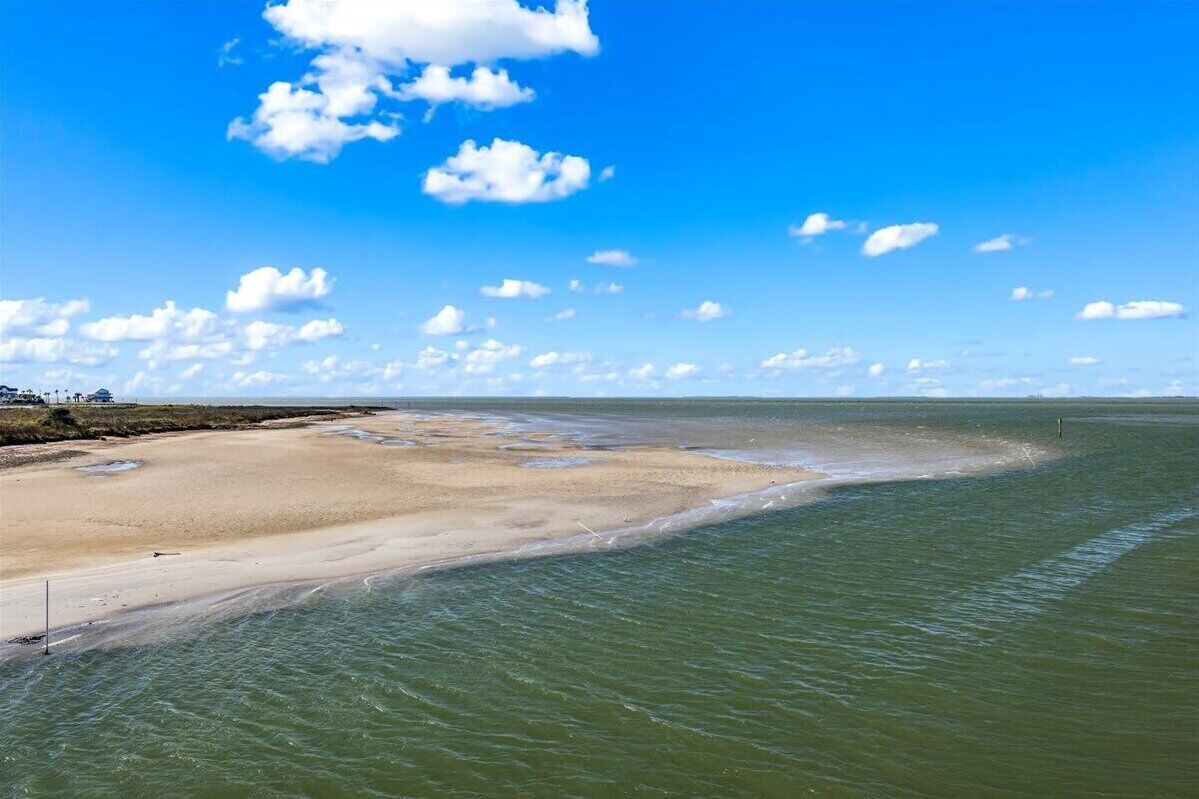 A coastal landscape with a sandy beach, calm water, and a clear blue sky with scattered clouds.