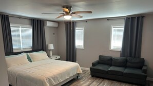 Een slaapkamer met een bed, een nachtkastje, een plafondventilator, een raam met jaloezieën en een grijze bank.