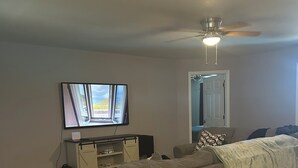 Een woonkamer met een houten eettafel, een grijze bank, een televisie en een plafondventilator.